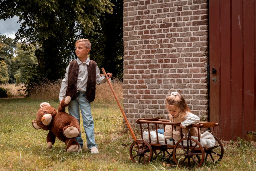 Kerstfoto waarbij een meisje in een oude houten bolderkar zit en haar broer de kar vooruit trekt. De jongen heeft een grote teddybeer vast. Beide zijn gekleed in bruine, winterse kledij.